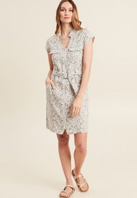 Abito a maniche corte color beige chiaro con motivo a foglie, zip frontale e cintura regolabile in vita; presenta due tasche frontali e una vestibilità comoda.