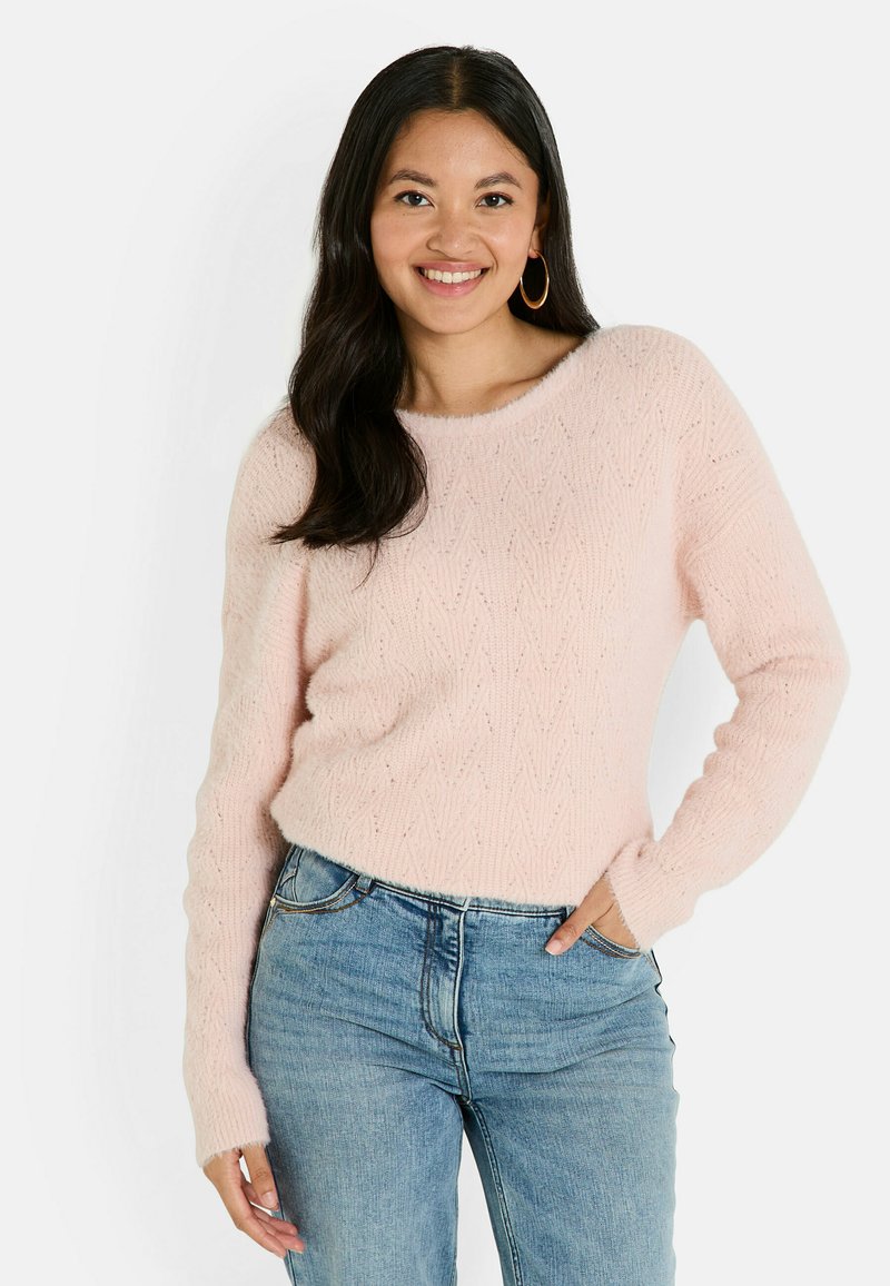 Pull en tricot rose clair avec un motif texturé, encolure ronde et manches longues, associé à un jean bleu. Tissu doux avec un aspect confortable.