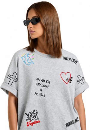 Femme aux cheveux bruns et raides portant des lunettes de soleil noires et un t-shirt gris avec diverses phrases imprimées et petits motifs graphiques.