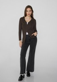 Cardigan marrón con escote en V y cierre de botones, combinado con jeans de pierna ancha negros. El modelo tiene el cabello largo y lleva zapatos negros.