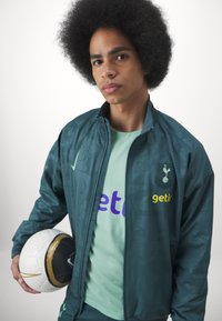 Giacca sportiva turchese con logo e motivi, indossata sopra una camicia verde chiaro con testo viola. Tienendo un pallone da calcio texturizzato abbinato.