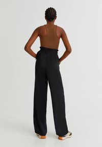 Personne avec des cheveux tressés portant un haut sans manches marron, un pantalon noir large taille haute et des baskets blanches, se tenant avec les mains sur les hanches.