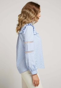 Chemise rayée bleu clair avec un col et des manches froncés, présentant des inserts en dentelle sur les manches, confectionnée en coton doux.