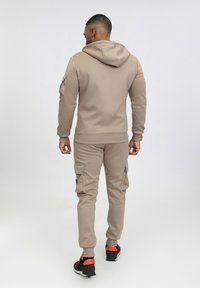 Sweatshirt beige à capuche et pantalon cargo assorti avec poches latérales, conçu dans un style ajusté. Porté avec des baskets noires et oranges.