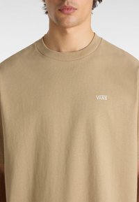 Béžové bavlněné tričko s kulatým výstřihem a malým bílým vyšívaným logem "VANS" na levé části hrudi. Hladká textura, volný střih.