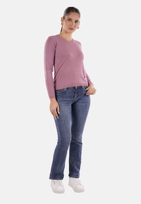 Maglione rosa a maniche lunghe con scollatura a V, design aderente, abbinato a jeans a campana blu e sneakers bianche, indossato da un modello su uno sfondo bianco.