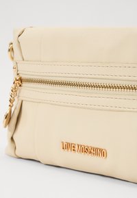 Beige nahast kott kuldsete detailidega, millel on tõmblukuga tasku, reljeefne logo ja tekstureeritud detailid külgedel.