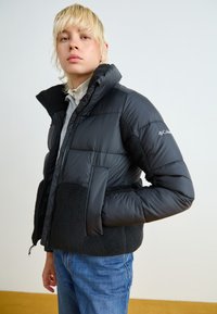 Svart pufferjakke med en pelsaktig nederdel, glidelås, høy krage og frontlommer, båret over en lysegrå topp og blå jeans.