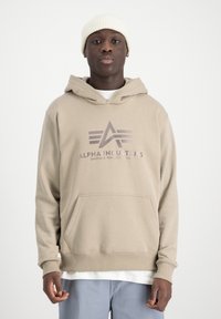 Sudadera con capucha beige y bolsillo canguro en la parte frontal, con un logo gráfico gris y puños de canalé. Llevada sobre una camiseta blanca.