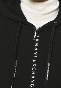 Felpa nera con zip che presenta un logo bianco lungo la zip, tessuto texturizzato e cordini regolabili. Indossata sopra un dolcevita bianco.