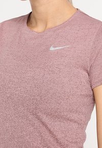 Camiseta de cuello redondo de color rosa melange con una sutil textura, que presenta un pequeño logo plateado de Nike en el lado izquierdo del pecho. Mangas cortas y tejido suave.