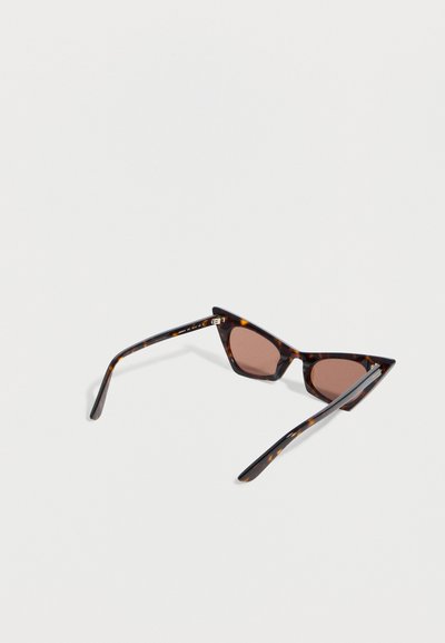 Γυαλιά ηλίου με σκελετό τύπου "γατόμματο" σε σχέδιο χελώνας και καφέ φακούς, που ακουμπούν σε μια επίπεδη ανοιχτογκρι επιφάνεια, σε γωνία που δείχνει το εμπρόσθιο μέρος και τα πλευρικά μπράτσα.