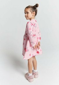 Junges Mädchen mit einem Haarknoten, das ein rosa Kleid mit Cartoon-Druck, weiße Socken und rosa flauschige Schuhe trägt, steht mit den Händen hinter dem Rücken.
