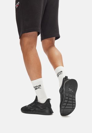 Gambe con pantaloncini neri Reebok, calze bianche Reebok e scarpe da ginnastica nere Reebok, che mostrano la suola e la vista laterale su sfondo bianco.