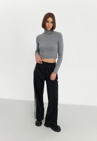 Un crop top gris à col roulé côtelé associé à un pantalon de sport noir avec des rayures blanches sur les côtés et un logo, ainsi qu'à des chaussures noires épaisses.