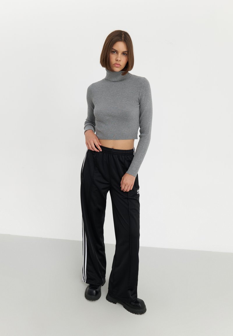 Un crop top gris à col roulé côtelé associé à un pantalon de sport noir avec des rayures blanches sur les côtés et un logo, ainsi qu'à des chaussures noires épaisses.