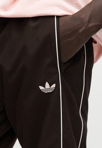 Καφέ αθλητικό παντελόνι με λευκές πλευρικές ρίγες, με ροζ κεντημένο λογότυπο Adidas trefoil στο αριστερό πόδι. Λεπτομέρεια φερμουάρ στην τσέπη.