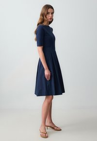 Navyblauwe jurk, getailleerde lijf, korte mouwen, geplooide rok. Gladde stof met een strak ontwerp en subtiele stikdetails.