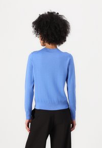 Sweater azul tricotado com mangas longas, decote redondo e bainha canelada. Textura suave e design ajustado. Usado com calças pretas.