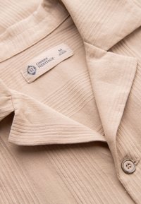 Gros plan sur le col d'une chemise en tissu beige texturé avec un bouton visible et une étiquette indiquant Ombre Heritage taille medium.