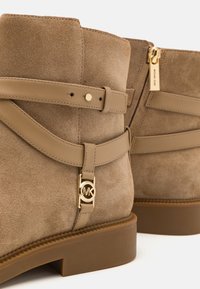 Bottines en daim marron clair avec des sangles en cuir assorties, détail du logo MK doré et tirette de fermeture éclair dorée sur semelle en caoutchouc lisse.