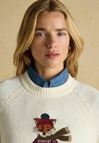 Krēmveida džemperis ar ribotu apkakli, kurā ir izšūts lapsa ar cepuri un šalli. Zem tā ir redzama zila džinsaina krekls.