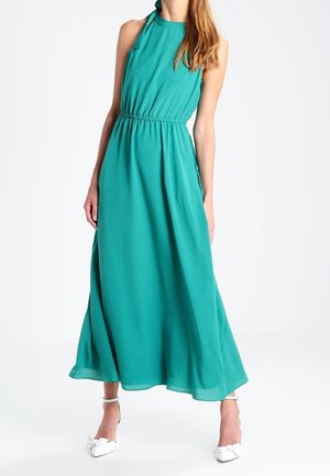 Femme portant une robe longue sans manches couleur sarcelle avec une taille froncée et des chaussures blanches à talons, se tenant devant un fond blanc uni.