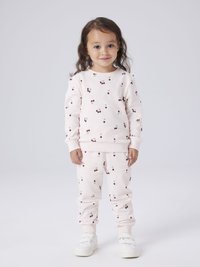 Jeune enfant aux longs cheveux bruns portant un survêtement rose clair à motifs cerises et des baskets blanches à scratch, debout devant un fond uni.