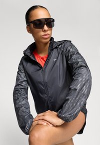 Zwarte sportjack met gestippelde mouwen, ritsvoorkant en capuchon. Draagt over een rode top. Donkere zonnebril maakt de atletische look compleet.