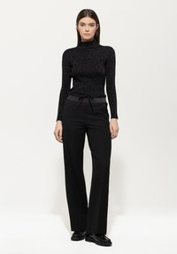 Schwarzes, tailliertes Rollkragenoberteil mit glitzernder Textur, kombiniert mit hochgeschnittenen, weiten schwarzen Hosen mit Kordelzug. 