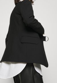 Blazer noir à la coupe structurée, avec une poche unique et un détail en métal argenté. Porté sur une chemise blanche à ourlet arrondi.