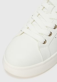 Sneaker sintetica bianca con punta arrotondata, lacci piatti e occhielli color oro. Presenta una texture liscia e una suola in gomma.