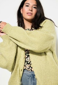 Cárdigan de punto verde claro con corte holgado, puños de canalé y botones grandes, superpuesto a una blusa negra con estampado floral.