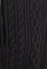 Dunkler gestrickter Pullover mit strukturierten Zopfmustern und einem Reißverschluss auf der linken Seite. Das Material wirkt weich und dick.