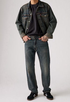 Chaqueta vaquera de color azul desgastado, camiseta oscura, vaqueros azules descoloridos y zapatos negros con rayas blancas. Ajuste casual con detalles de costuras visibles.