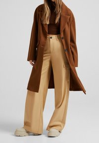 Długi brązowy płaszcz na prążkowanym brązowym golfie, zestawiony z szerokimi beżowymi spodniami i masywnymi beżowymi butami na platformie. Gładkie tekstury i dopasowany krój.