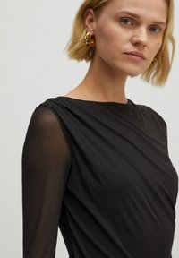 Schwarzes Langarmoberteil mit einer drapierten Gestaltung, transparentem Material an den Ärmeln und einer glatten Silhouette. Beinhaltet minimale Details am Ausschnitt.