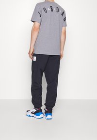 Grå kortärmad T-shirt med "JORDAN" i svarta bokstäver på ryggen, kombinerad med löst sittande marinblå byxor och vita, svarta och blå sneakers.