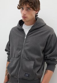 Felpa grigia con zip, dotata di tasche a marsupio, polsini a coste e una texture morbida. Presenta un'etichetta con logo sul lato sinistro anteriore in basso.