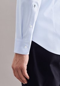 Camicetta azzurra chiara con una texture liscia, chiusure a bottone sui polsini e un dettaglio ricamato a forma di rosa vicino alla spalla.
