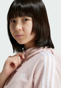 Sudadera con cremallera de color rosa claro con acentos de tres rayas blancas en los hombros. Presenta un pequeño logo blanco de Adidas en el pecho. Material suave, cuello redondeado.