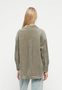 Oliwkowa koszula z kordurojowego materiału z detalem wzdłuż z tylu, zapinanymi mankietami i luźnym krojem. Tkanina ma miękką fakturę.