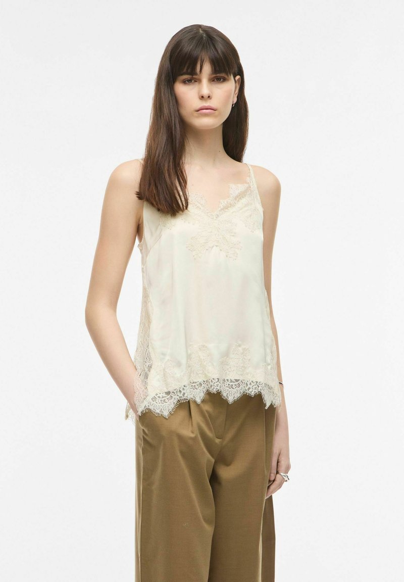 Jeune femme aux longs cheveux foncés portant un haut sans manches crème orné de dentelle et un pantalon brun ample, debout avec une main dans la poche.