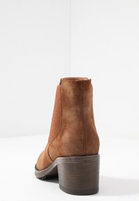Brown suede ankle boot with a textured elastic side and a stacked wooden heel. The back features a curved design and contrasting stitching.
