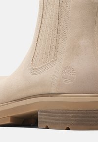 Beige Wildlederstiefel mit vertikalen Nähten, geprägtem Logo und einem gestapelten Holzabsatz mit strukturiertem Oberflächen.