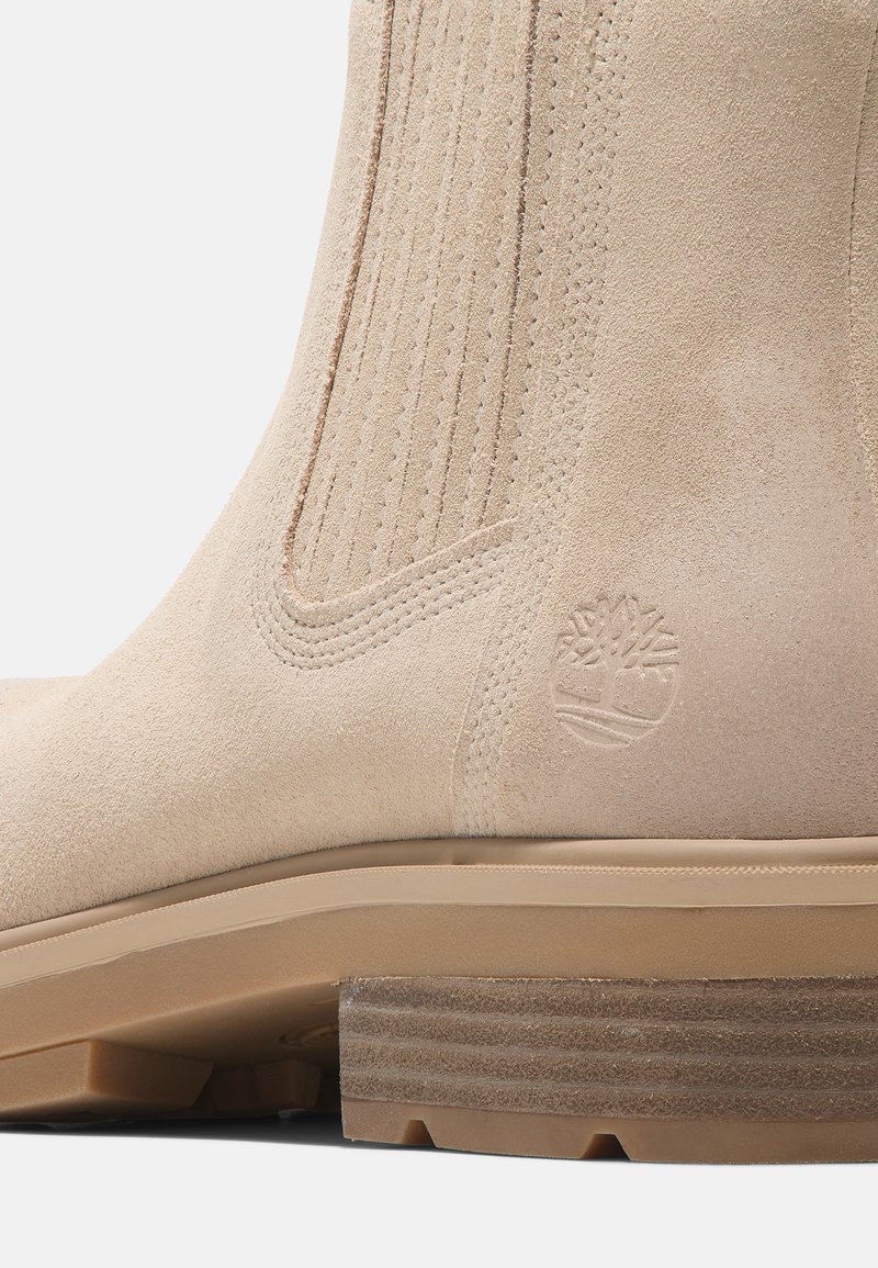 Botte en suède beige avec des coutures verticales, un logo en relief et un talon en bois empilé à la surface texturée.