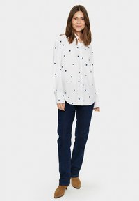 White button-up shirt with small blue heart patterns, paired with dark blue jeans and tan ankle boots, in a standing pose on a neutral background.