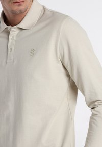 Beige Langarm-Polohemd aus strukturiertem Stoff mit einer Drei-Knopf-Leiste und einem dezenten gestickten Logo auf der Brust.