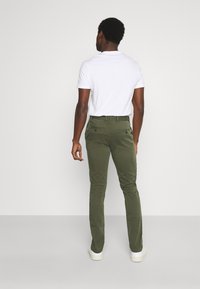 Olijfgroene chino's met een slim fit, gemaakt van stof met een gladde textuur. Bevat twee achterzakken met knoopsluitingen. Draagt met een wit t-shirt.