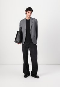 Man wearing gray pinstripe blazer, black shirt and pants, holding large black leather tote bag against plain white background.